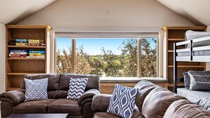 Una habitación acogedora con un sofá de cuero marrón, una ventana con vistas a árboles y una estantería con juegos de mesa.