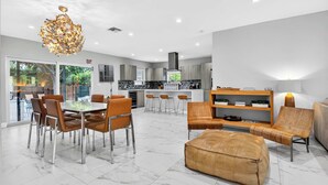 Una cocina moderna con estufa integrada, un comedor con mesa y sillas, y una sala de estar con sofá y reposapiés.
