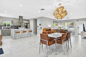 Une cuisine moderne dotée d’un îlot central, un coin repas avec une table blanche et des chaises marron, et un espace de détente avec un canapé et un téléviseur.