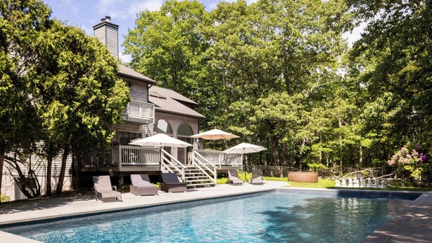 Une piscine entourée d’arbres, avec des chaises longues et des parasols, et une maison dotée d’un balcon.