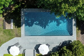 An aerial view of a rectangular swimming pool with clear blue water, surrounded by a paved deck and two white umbrellas.
