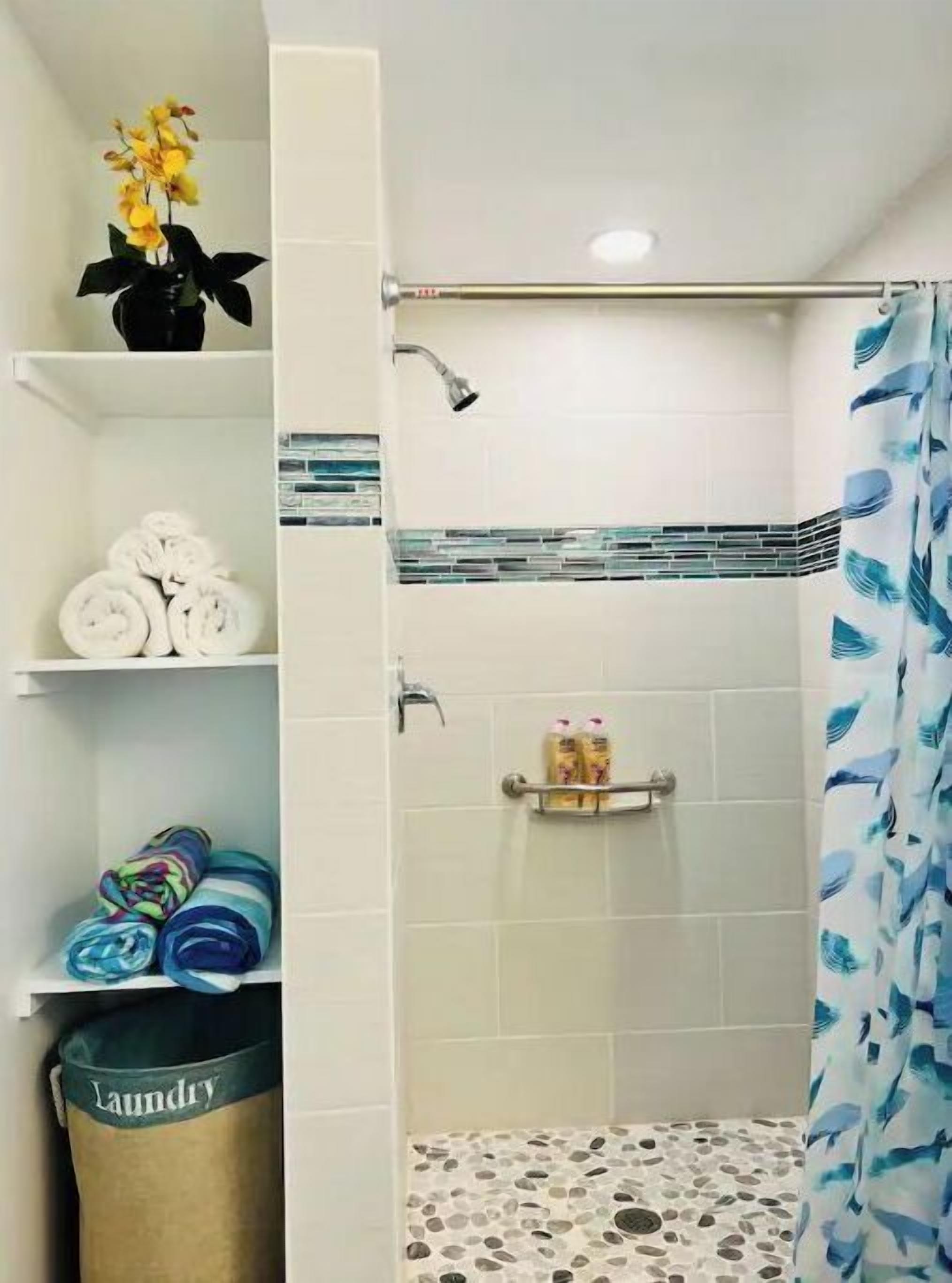 A bathroom with a shower, a towel rack, a shelf with folded towels, a laundry basket, and a potted plant.