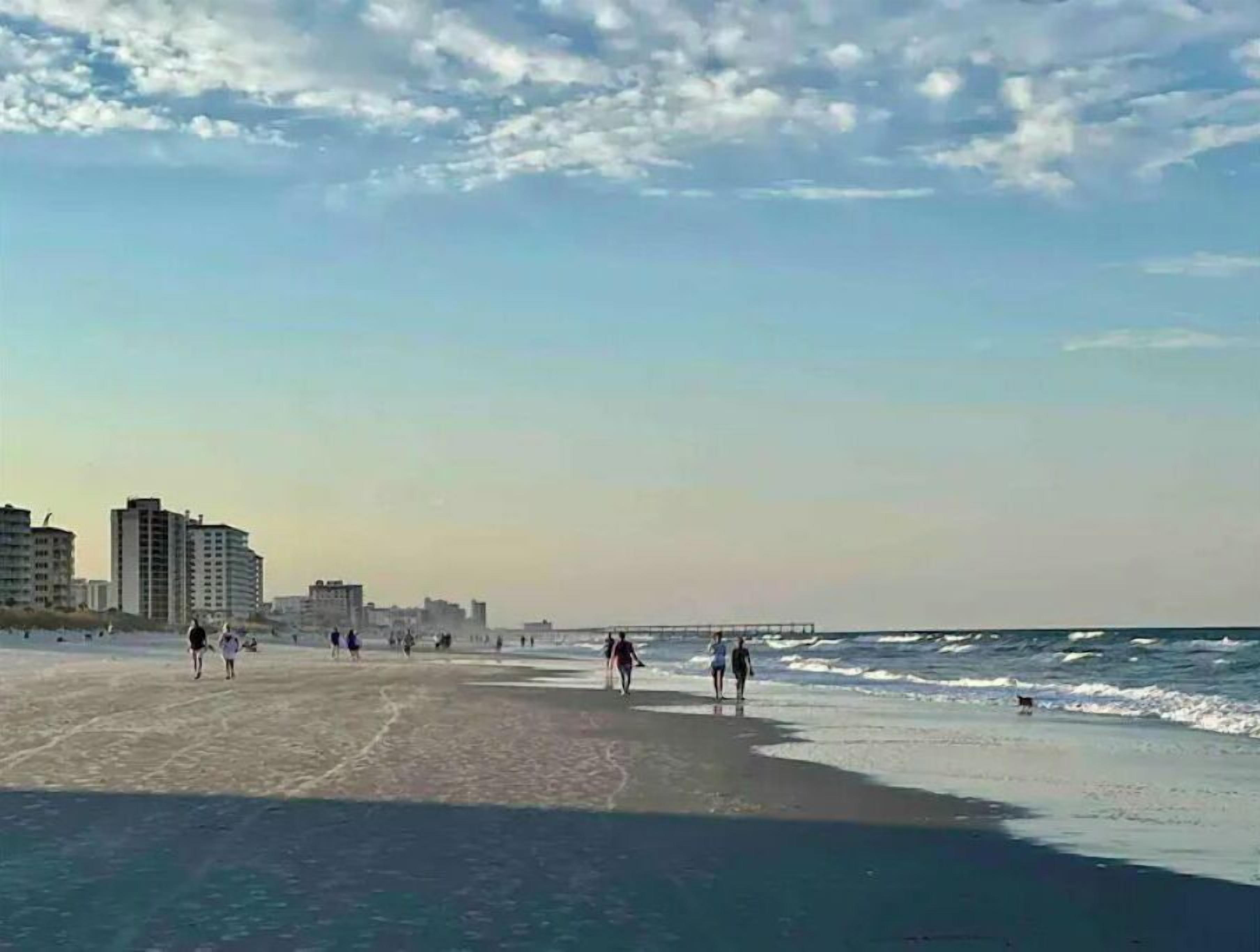 Una playa con gente caminando, edificios al fondo y un cielo despejado.