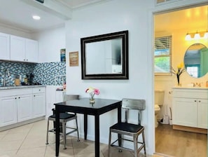 Compact kitchen with white cabinetry, black dining table, and adjacent bathroom with vanity and mirror.