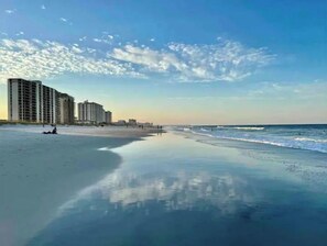 Uma praia com céu claro, mar calmo e edifícios altos ao fundo.