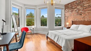 Ein Schlafzimmer mit einem großen Bett, einem Schreibtisch mit Stuhl und Blick auf Grün hinter großen Fenstern.
