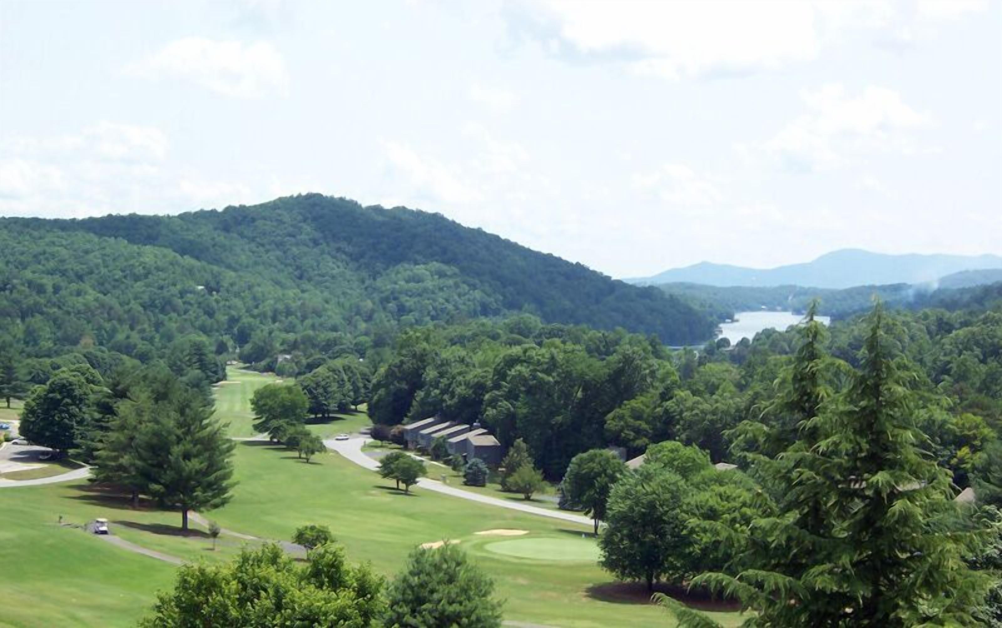 Un campo de golf con club house, rodeado de un bosque denso y colinas al fondo.