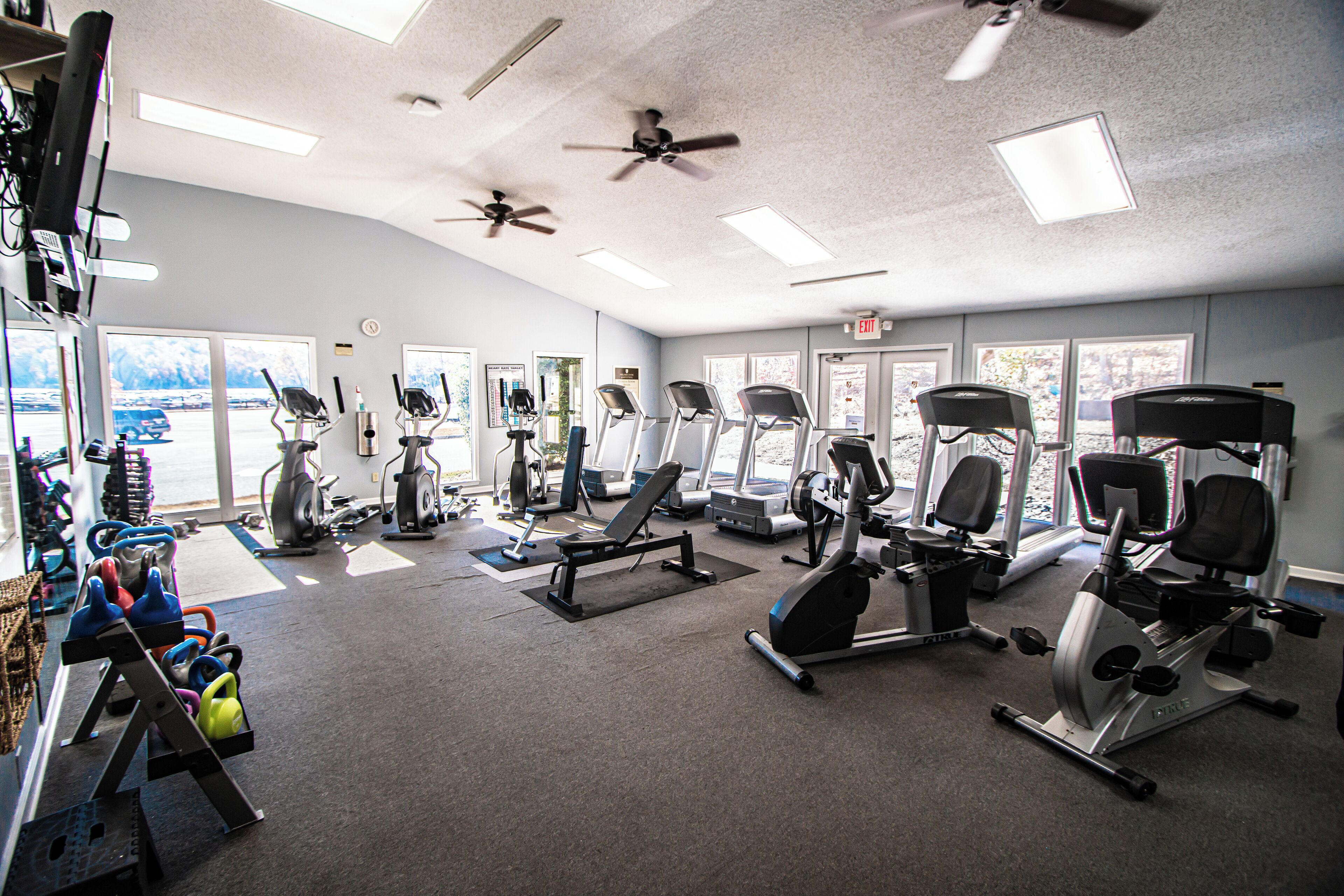 A gym with various exercise equipment, ceiling fans, and large windows.