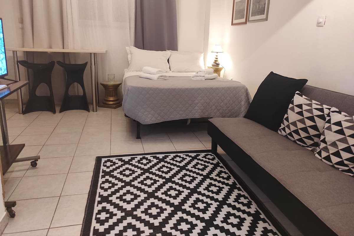 A hotel room with a bed, a sofa, a desk with a chair, a small table with a lamp, and a patterned rug.