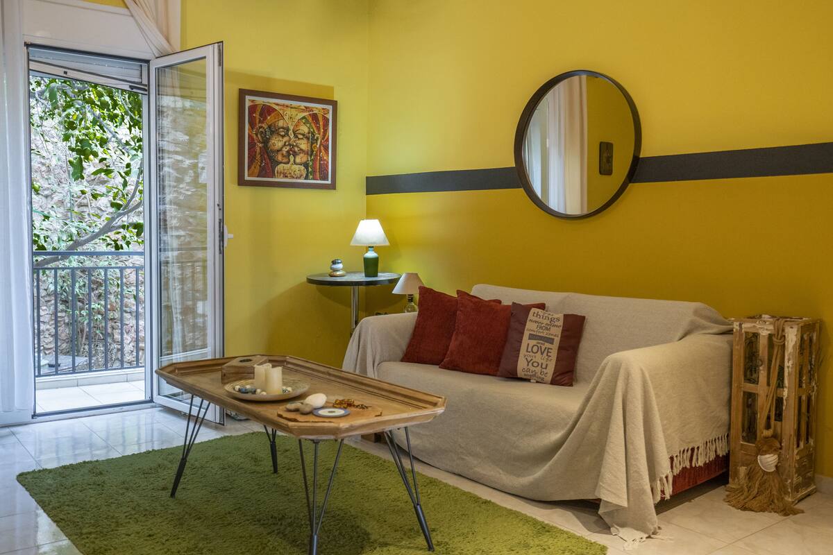 A room with a green rug, a sofa with red cushions, a round mirror, and a small table with a lamp and decorative items.