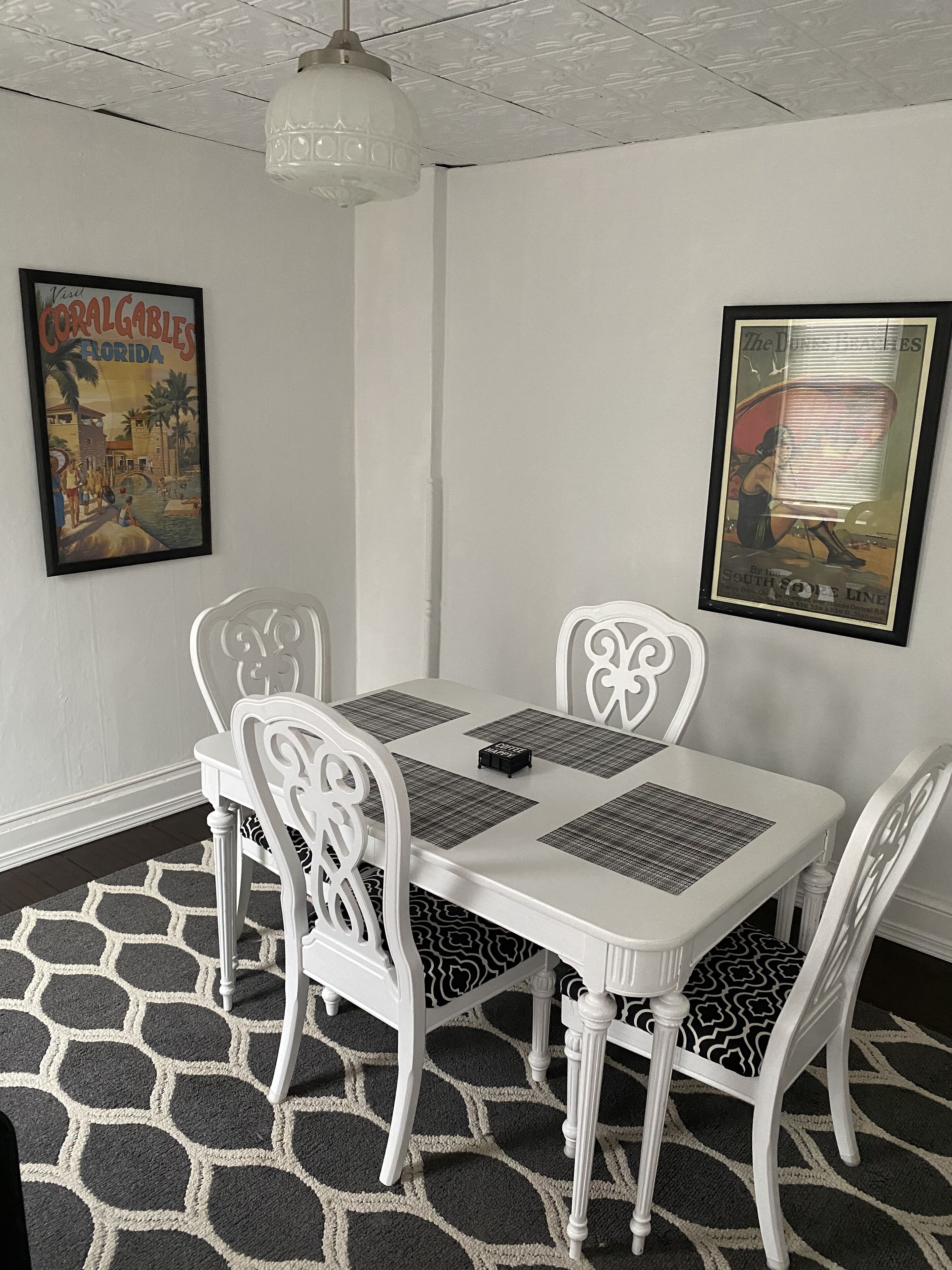 Un comedor con una mesa y sillas blancas, un tapete estampado y dos carteles enmarcados en la pared.