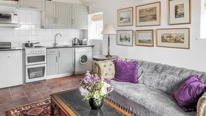 Une cuisine avec des meubles blancs, une cuisinière et un lave-linge. On y trouve un canapé gris avec des coussins violets et un vase de fleurs.