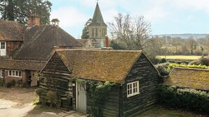 Une maison de campagne traditionnelle en bois, dotée d’un petit porche, avec une église en pierre en arrière-plan et un ciel dégagé.