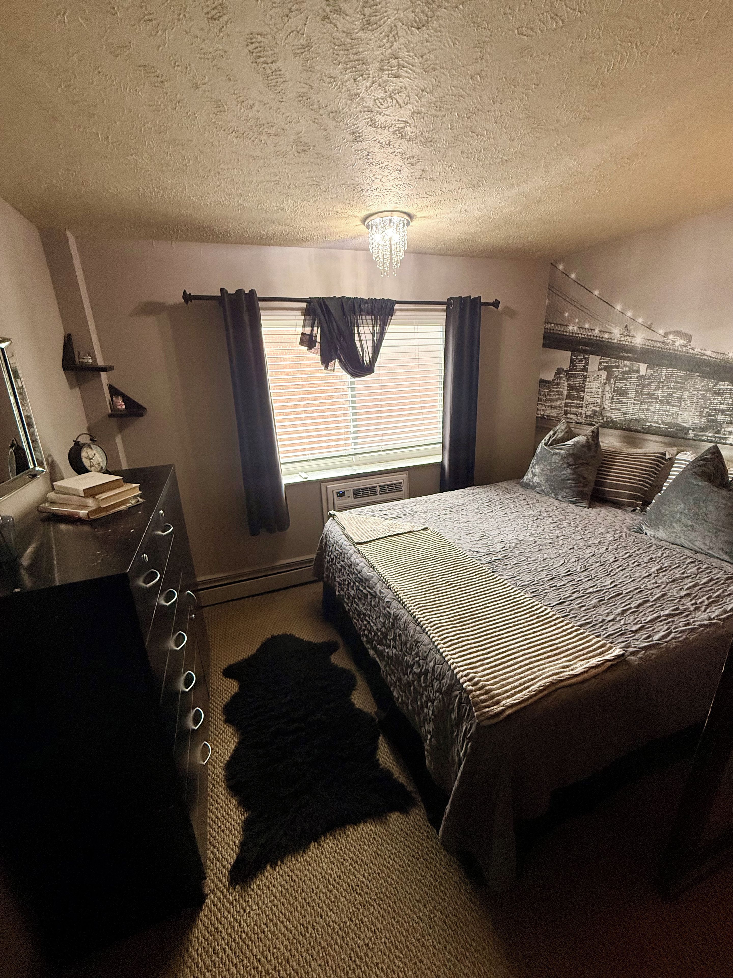 Un dormitorio con cama, cómoda, espejo, ventana con cortinas y vistas a la ciudad.
