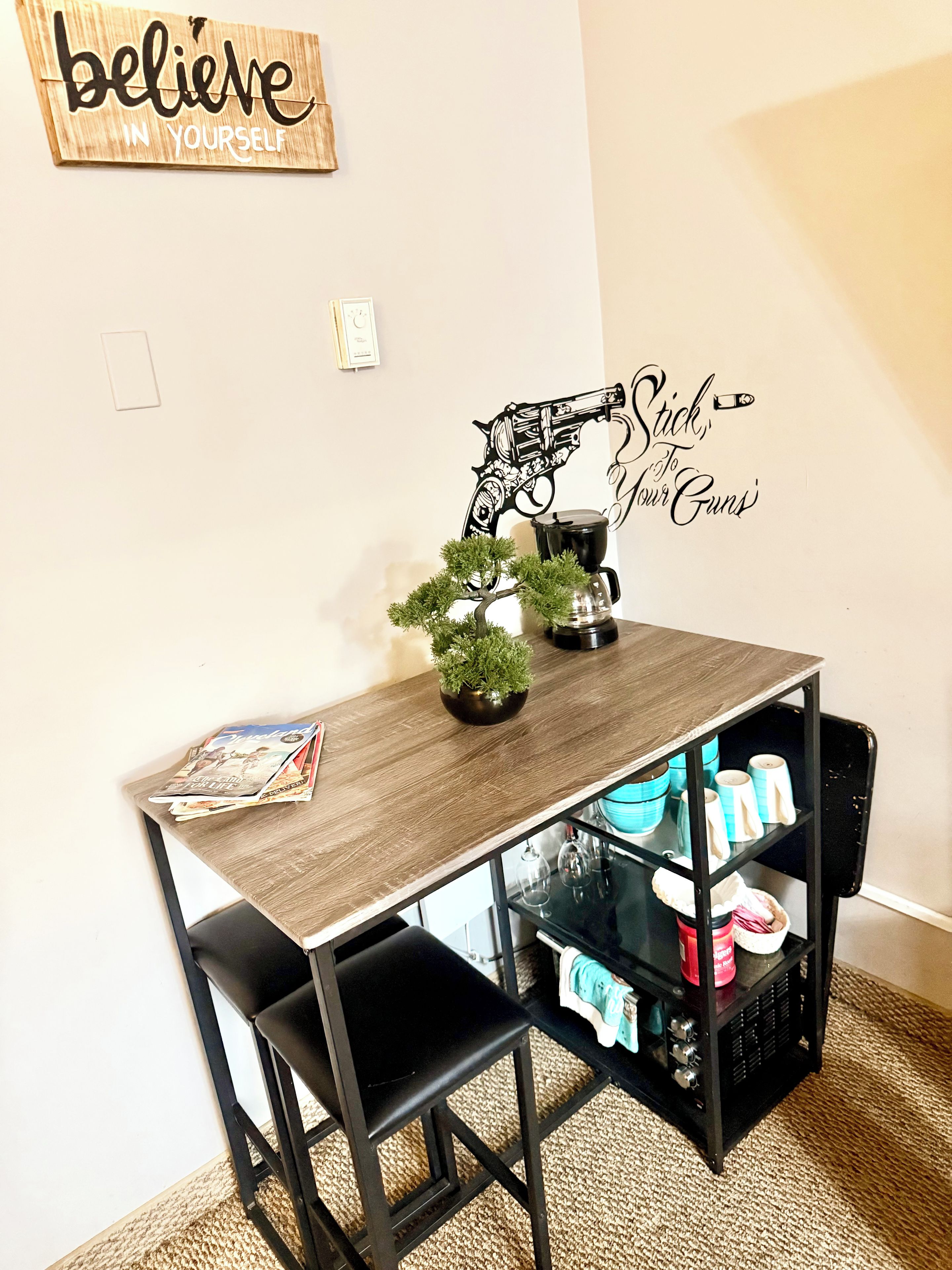 Comedor con mesa de madera, sillas negras, cafetera, planta en maceta y un adorno en la pared.