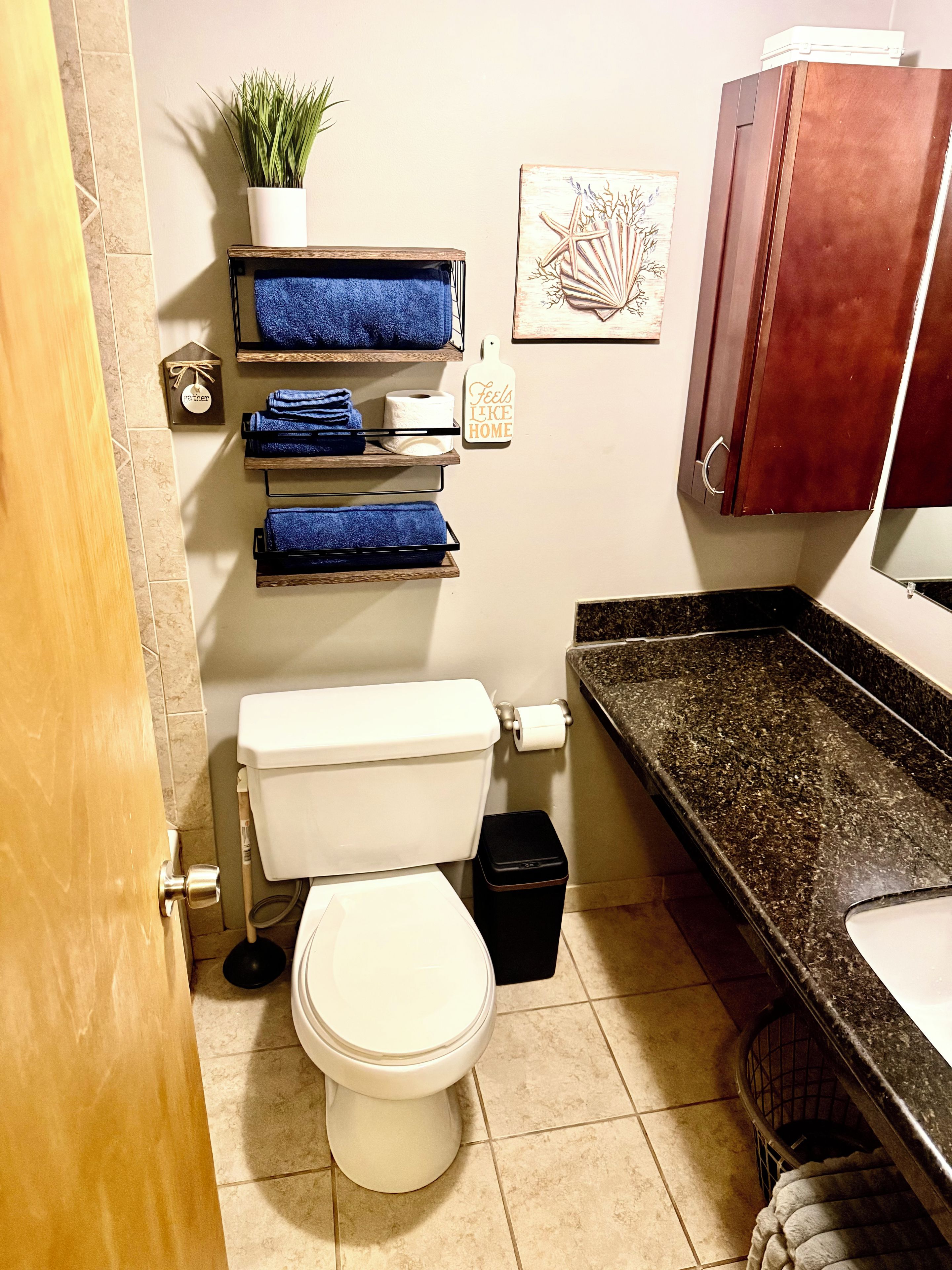 A bathroom with a toilet, a sink, a trash bin, a towel rack with towels, a potted plant, and a decorative wall piece.