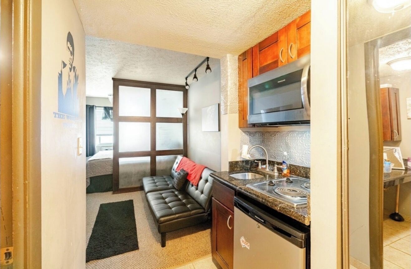 Una cocina compacta con microondas, estufa y lavamanos. Hay un sofá de cuero negro y una alfombra pequeña en la sala.