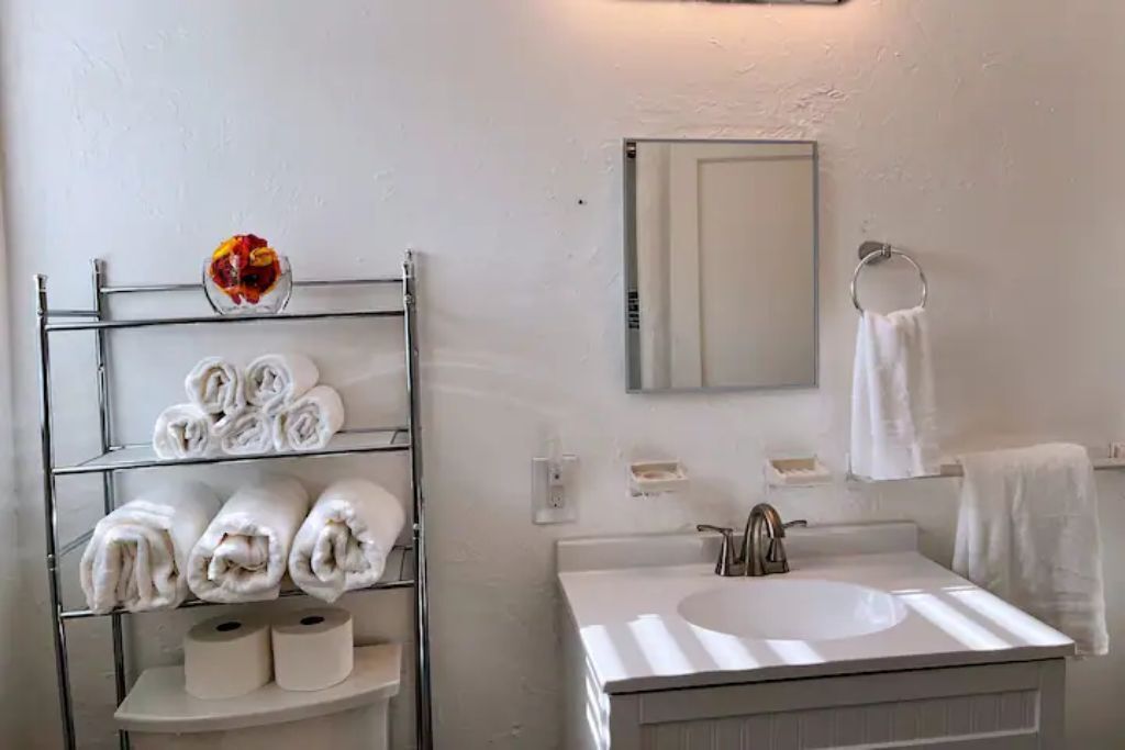 A bathroom with a white sink, a mirror, a towel rack, and a shelf with towels and toilet paper.
