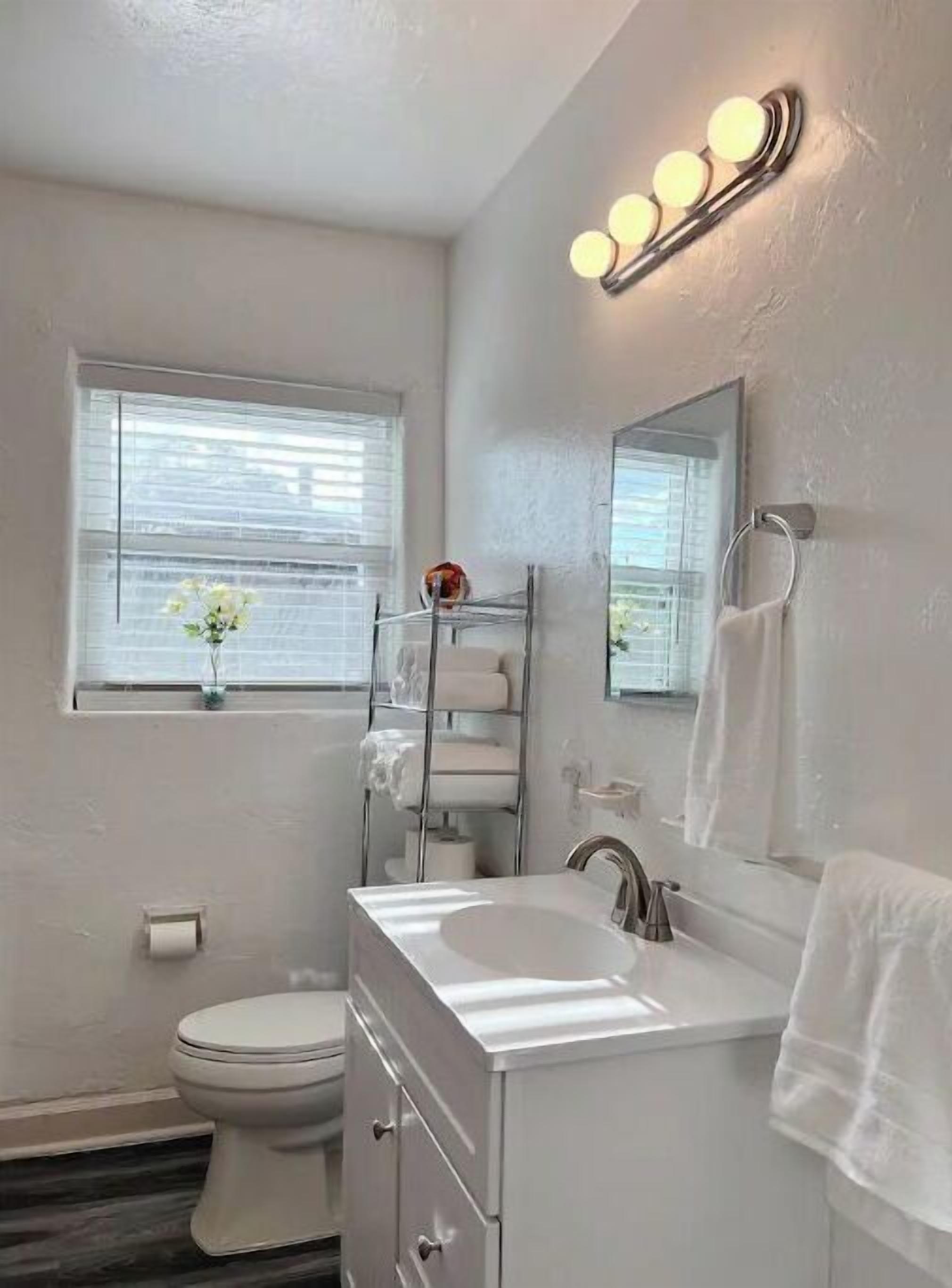 A modern bathroom with a toilet, sink, and mirror. There is a window with blinds and a towel rack.