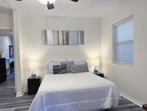 A bedroom with a bed, two bedside tables with lamps, a ceiling light, a window with blinds, and a wall with decorative panels.