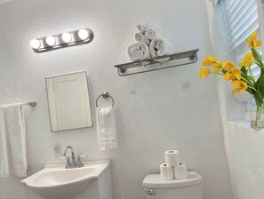 A bathroom with a sink, mirror, towel rack, toilet, and a shelf with rolled towels and toilet paper.