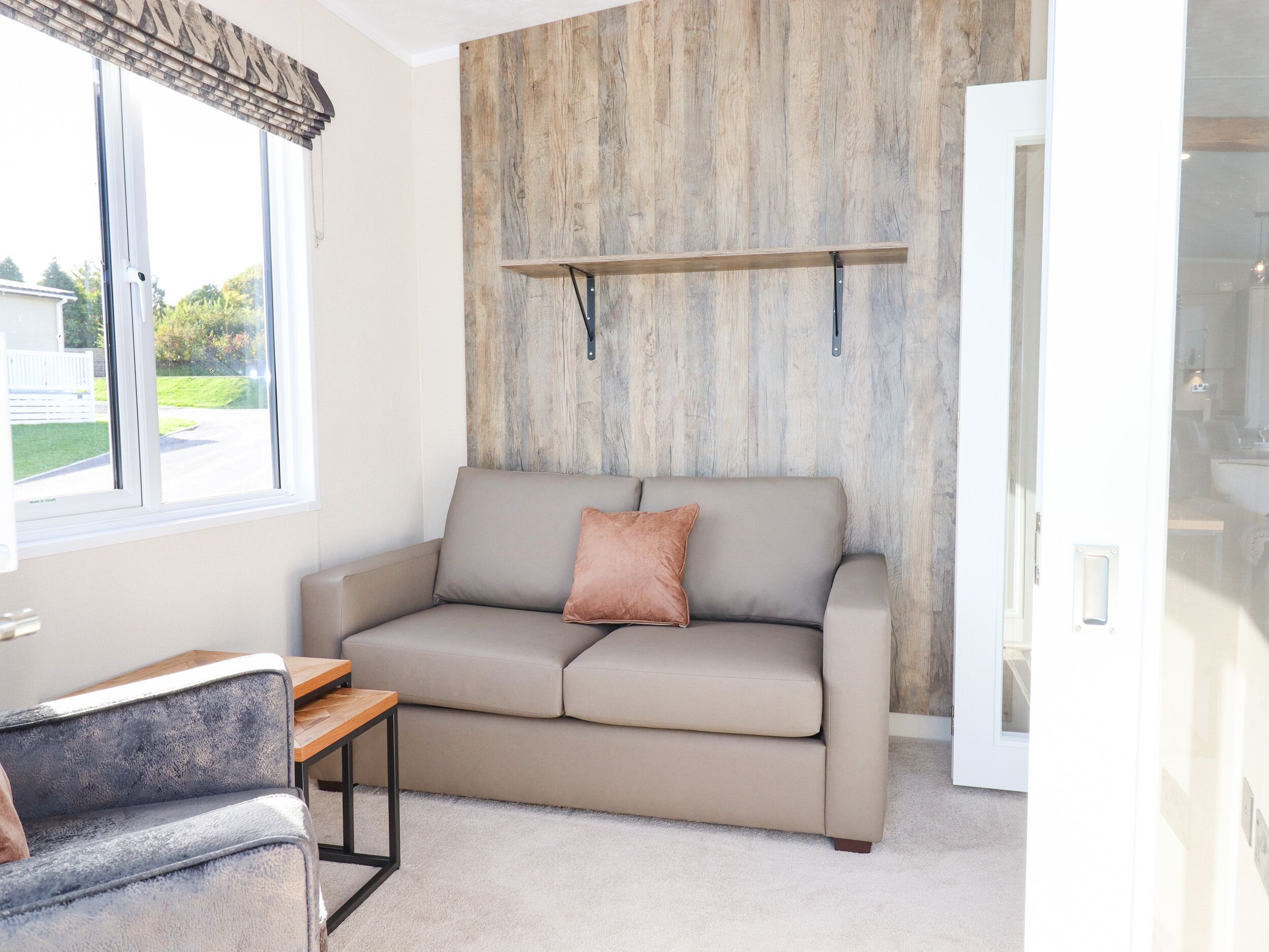 Una sala de estar moderna con un sofá color beige, una mesita de madera y un ventanal con vistas a una casa y vegetación.