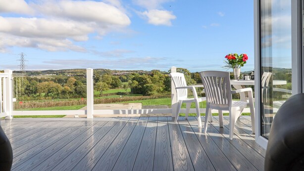 Une terrasse en bois, avec des chaises blanches et une table, d’où l’on peut admirer un paysage pittoresque composé de collines et d’un pylône de ligne électrique.
