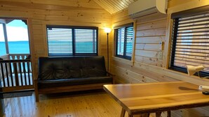 A wooden cabin interior with a sofa, a dining table, and a view of the ocean through large windows.