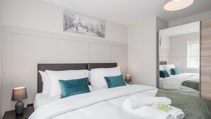 A neatly made bed with white bedding and teal pillows, a bedside lamp, and a painting of a cityscape on the wall.