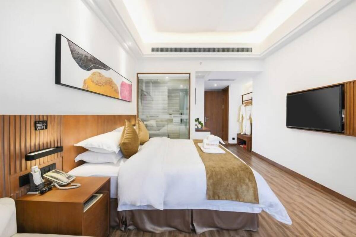 A hotel room with a bed, bedside table with a phone, a TV on the wall, and a painting above the bed.