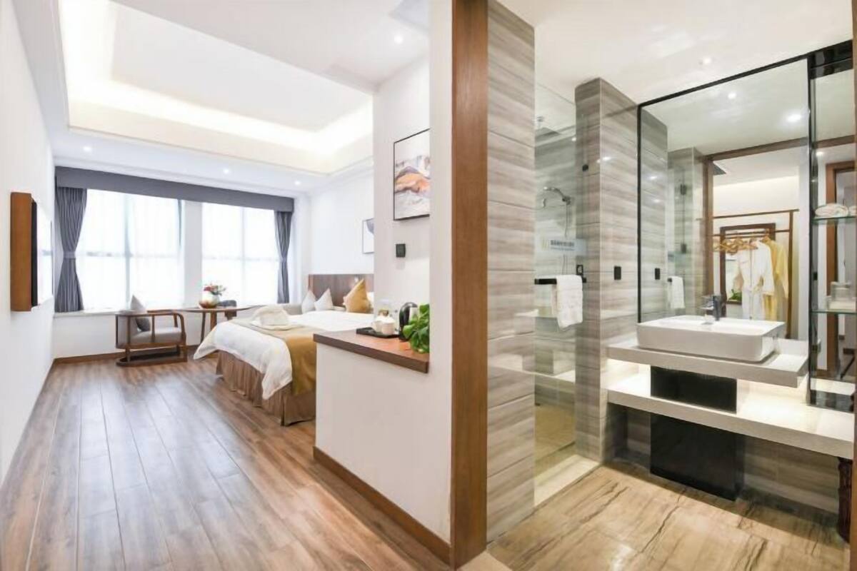 A modern hotel room with a wooden floor, a bed, a desk, a large window, and a bathroom with a glass shower enclosure.