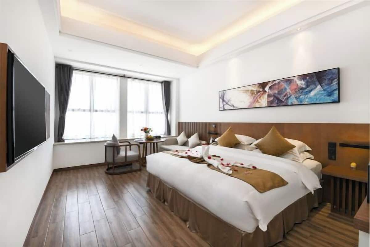 A hotel room with a large bed, a flat-screen TV, a window with curtains, a painting on the wall, and a small table with a vase.