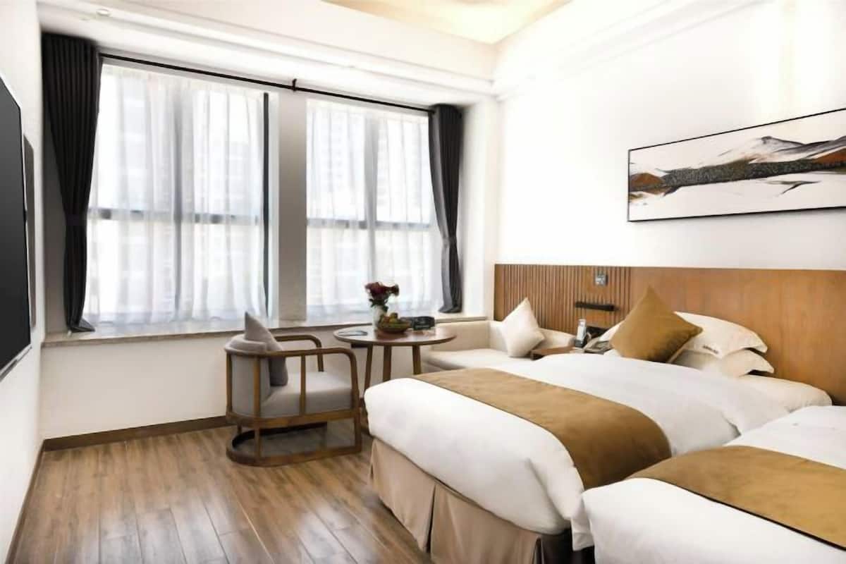 A hotel room with two beds, a small round table with a vase, a chair, and a window with curtains.