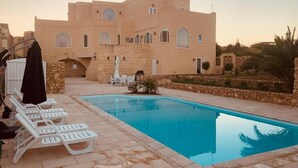 Un espace piscine avec des chaises longues et un bâtiment doté de fenêtres en arc et de murs en pierre.