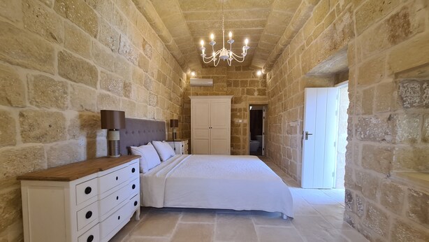 Une chambre aux murs de pierre, avec un lit, une commode et un lustre.