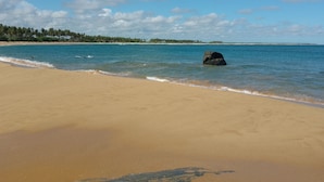 Una playa con agua azul cristalina, una roca grande en el centro y una costa lejana con árboles.