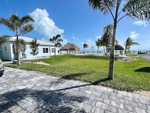 Um resort com um caminho pavimentado, palmeiras, uma construção com telhado de palha e vista para o oceano.