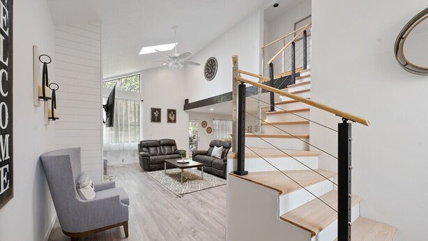Un salon spacieux avec un fauteuil gris, un canapé et une table basse. Un escalier avec une rampe en bois mène à l’étage supérieur.
