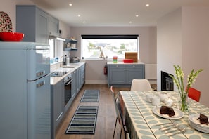A modern kitchen with grey cabinets, a red bowl, and a dining area with a table set for two. - Stone’s Throw: A pet-friendly holiday bungalow in Rhosneigr (Rhosneigr)