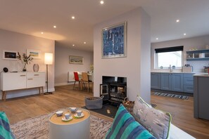 A modern living room with a fireplace, a dining area, and a kitchen with blue cabinets. - Stone’s Throw : Pet-Friendly 4‑Bed Bungalow Near Beach in Rhosneigr | Managed by Anglesey Holiday Le (Rhosneigr)