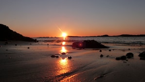 Uma praia com o sol se pondo sobre a água, projetando um reflexo na areia molhada.
