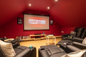 A home theater with a large screen displaying 'MARVEL STUDIOS,' black leather recliners, a wooden coffee table, and a bookshelf with DVDs.
