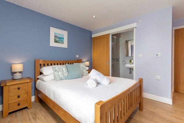 A bedroom with a wooden bed, a nightstand with drawers, a framed picture on the wall, and a bathroom visible through an open door.