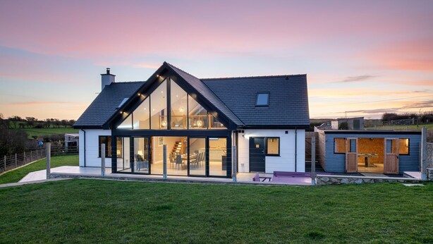 Une maison moderne à deux étages, dotée de grandes fenêtres en verre, d’un toit en pente et d’une pelouse bien entretenue.