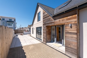 A modern house with a wooden exterior, a tiled patio, and a sliding glass door leading to a covered outdoor area. - Trem Y Don: Modern 5‑Bed Beachside Retreat Sleeping 10 in Rhosneigr | Managed by Anglesey Holiday Le (Rhosneigr)