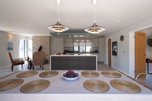 A modern kitchen with a central island, pendant lighting, and a dining area with a patterned rug and a bowl of grapes. - Trem Y Don: Modern 5‑Bed Beachside Retreat Sleeping 10 in Rhosneigr | Managed by Anglesey Holiday Le (Rhosneigr)