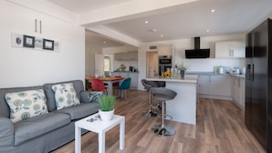 Een modern appartement met een grijze bank, een witte bijzettafel met een plant, een keuken met een zwarte televisie en een eethoek met rode stoelen.
