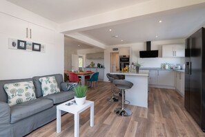 A modern living room with a grey sofa, a white coffee table, and a kitchen area with a black refrigerator, white cabinets, and a flat-screen TV.