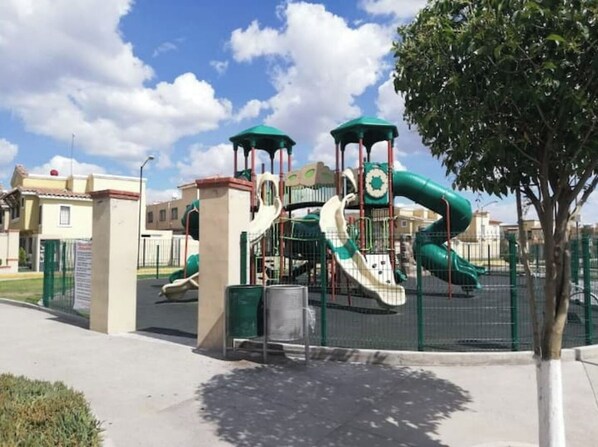 Kids can play on the playground's green slide and climbing structure, surrounded by a secure fence.