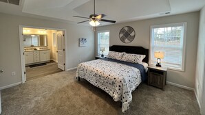 Ein Schlafzimmer mit Bett, Deckenventilator, Fenster mit Jalousien sowie Bad mit Spiegel und Waschbecken.